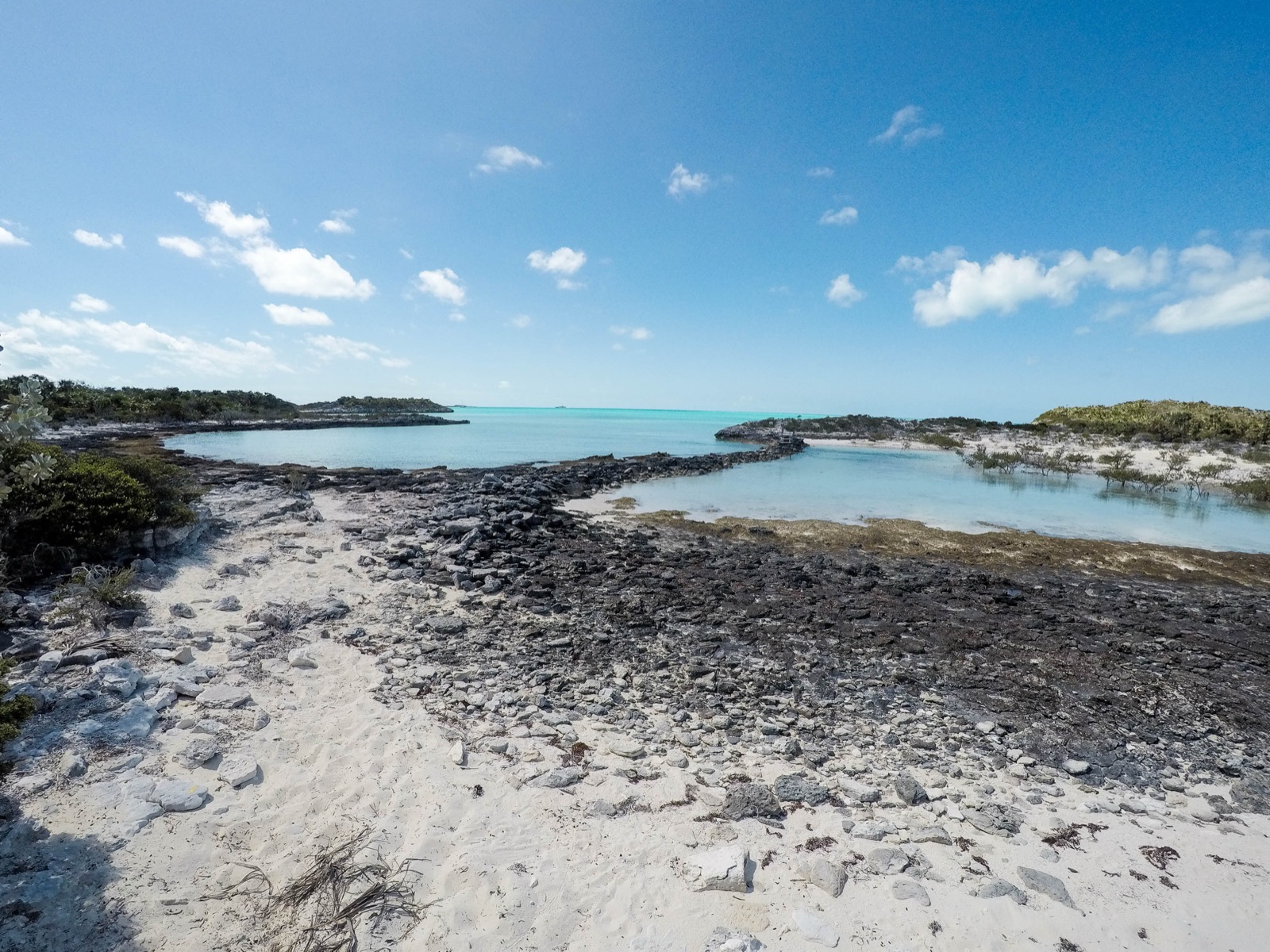 Rocky causeway between a turquoise bay and a sandy pond, with scrub and dunes beyond